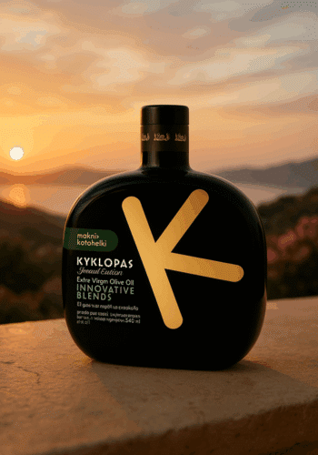 Schwarze, flache Flasche mit großem goldfarbenem „K“-Design und der Aufschrift „KYKLOPAS Special Edition Extra Virgin Olive Oil Innovative Blends, Makri-Kothelki“ steht auf einem Steinvorsprung. Im Hintergrund ein malerischer Sonnenuntergang über Meer und Hügeln.