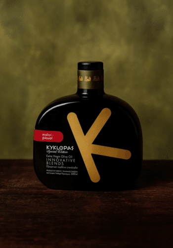 Schwarze, flache Flasche mit goldfarbenem „K“-Design und Aufschrift „KYKLOPAS Special Edition Extra Virgin Olive Oil Innovative Blends, Makri-Picual“ auf dunklem Holz vor verschwommenem, olivgrünem Hintergrund.